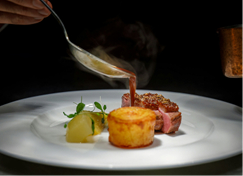 Um prato gourmet com um pedaço de carne assada, decorado com um molho sendo cuidadosamente derramado de uma colher. Ao lado, há uma porção de purê e um pequeno bolinho. O fundo é escuro, destacando a apresentação elegante da refeição.