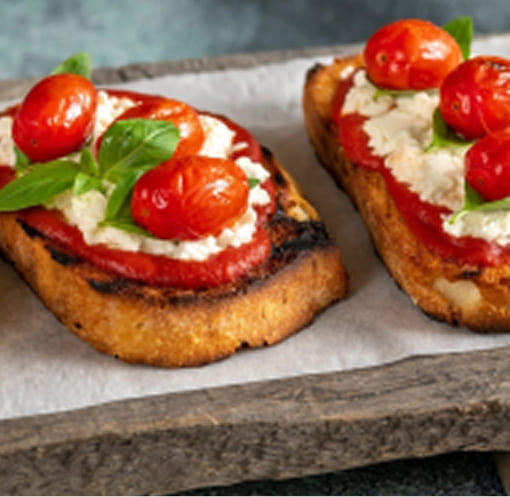 Duas torradas crocantes dispostas em uma tábua de madeira, cobertas com molho de tomate, queijo cremoso e tomates-cereja grelhados. Folhas de manjericão fresco decoram o prato, proporcionando uma apresentação colorida e apetitosa.